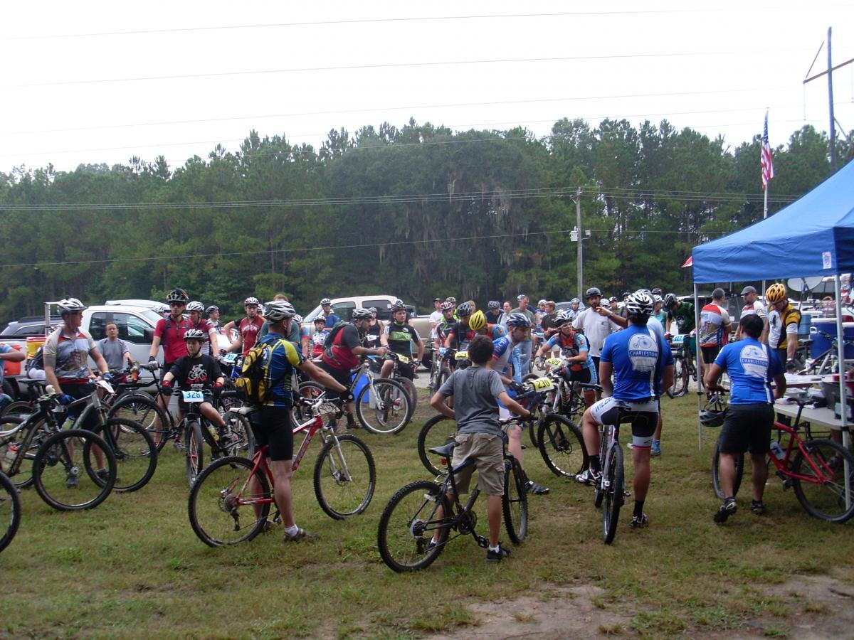 A large group of people gathered in a grassy area, preparing for a mountain biking event. Participants are chatting and adjusting their bicycles, with some wearing helmets and biking gear. A blue tent is set up nearby, and various vehicles are parked in the background. Trees line the area, creating a natural setting for the event. Wannamaker Park mountain bike trail.