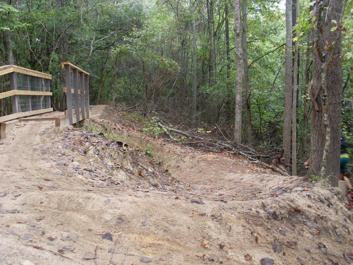 A winding dirt path through a wooded area, featuring a wooden bridge with low railings on the left. The landscape is surrounded by trees and underbrush, with fallen leaves scattered on the ground. Wannamaker Park mountain bike trail.