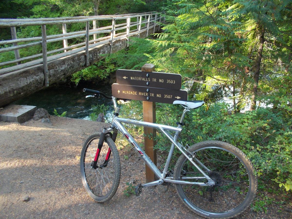 A silver mountain bike is parked on a dirt path next to a wooden bridge. Beside the bike, there are directional signs indicating the trails: "WATERFALLS TR NO 3503" and "MCKENZIE RIVER TR NO 3507." Lush green trees and a stream can be seen in the background, creating a natural, scenic environment. Mckenzie River Trail mountain bike trail.