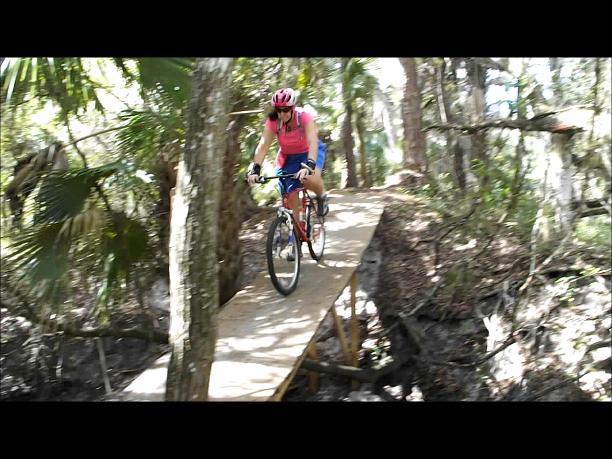 A mountain biker wearing a pink helmet and top rides along a narrow wooden bridge in a dense forest, surrounded by trees and greenery. Sunlight filters through the leaves, creating a natural and adventurous atmosphere. North Port Mountain Bike Trails mountain bike trail.