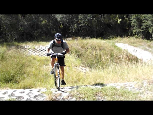 A mountain biker navigating a rocky path through a grassy area, wearing a helmet and a backpack, with trees in the background. The cyclist is focused on maintaining balance as they ride over uneven terrain. North Port Mountain Bike Trails mountain bike trail.