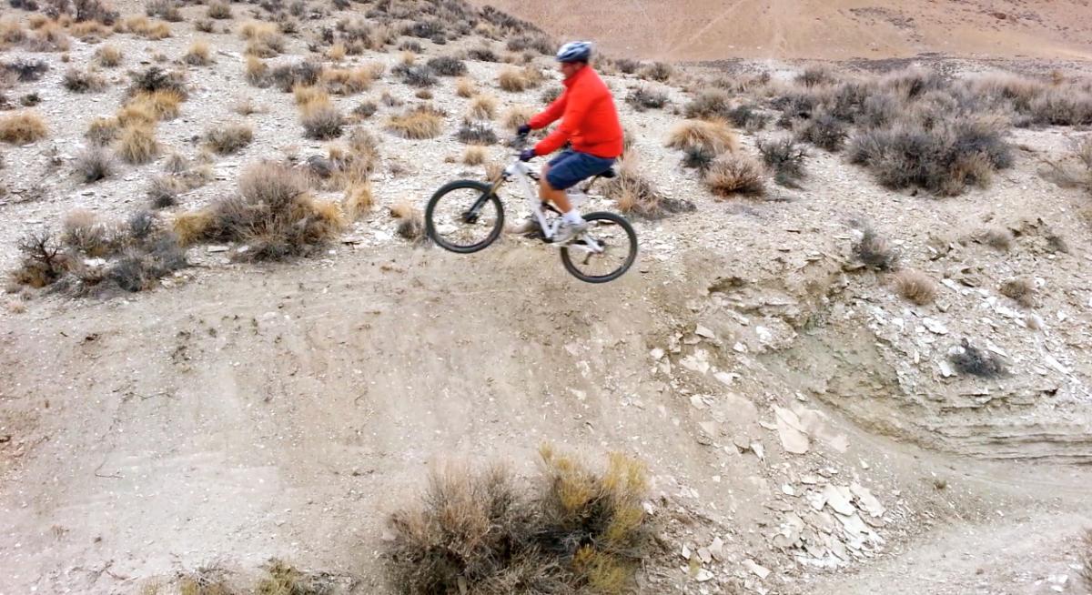 A person in a red jacket and shorts riding a mountain bike over a dirt ramp in a rugged outdoor landscape, surrounded by sparse vegetation and rocky terrain. Tnt mountain bike trail.