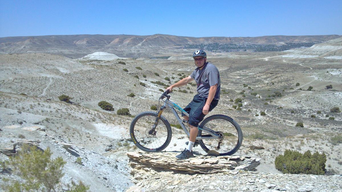 A person on a mountain bike stands on a rock outcrop overlooking a vast, arid landscape with rolling hills and sparse vegetation under a clear blue sky. Tnt mountain bike trail.