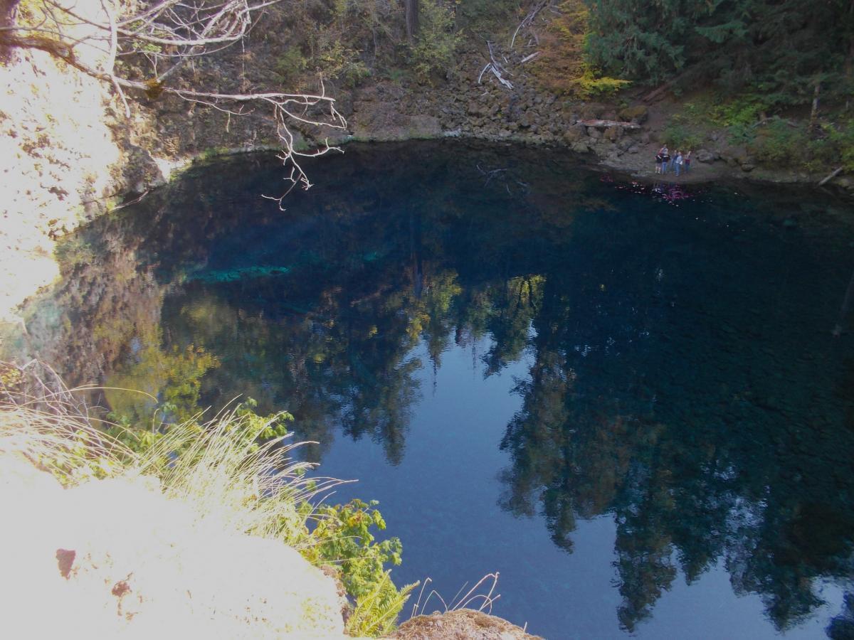 A tranquil, clear blue pond surrounded by lush greenery and rocky terrain, with reflections of trees in the water. A few people are seen at the water's edge, enjoying the scenery. Mckenzie River Trail mountain bike trail.