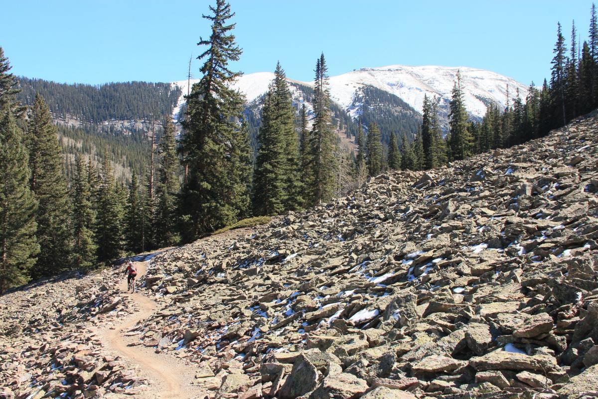 A hiker navigating a winding dirt trail through a rocky landscape, surrounded by tall evergreen trees and snow-capped mountains in the distance, under a clear blue sky. Prospect Trail mountain bike trail.