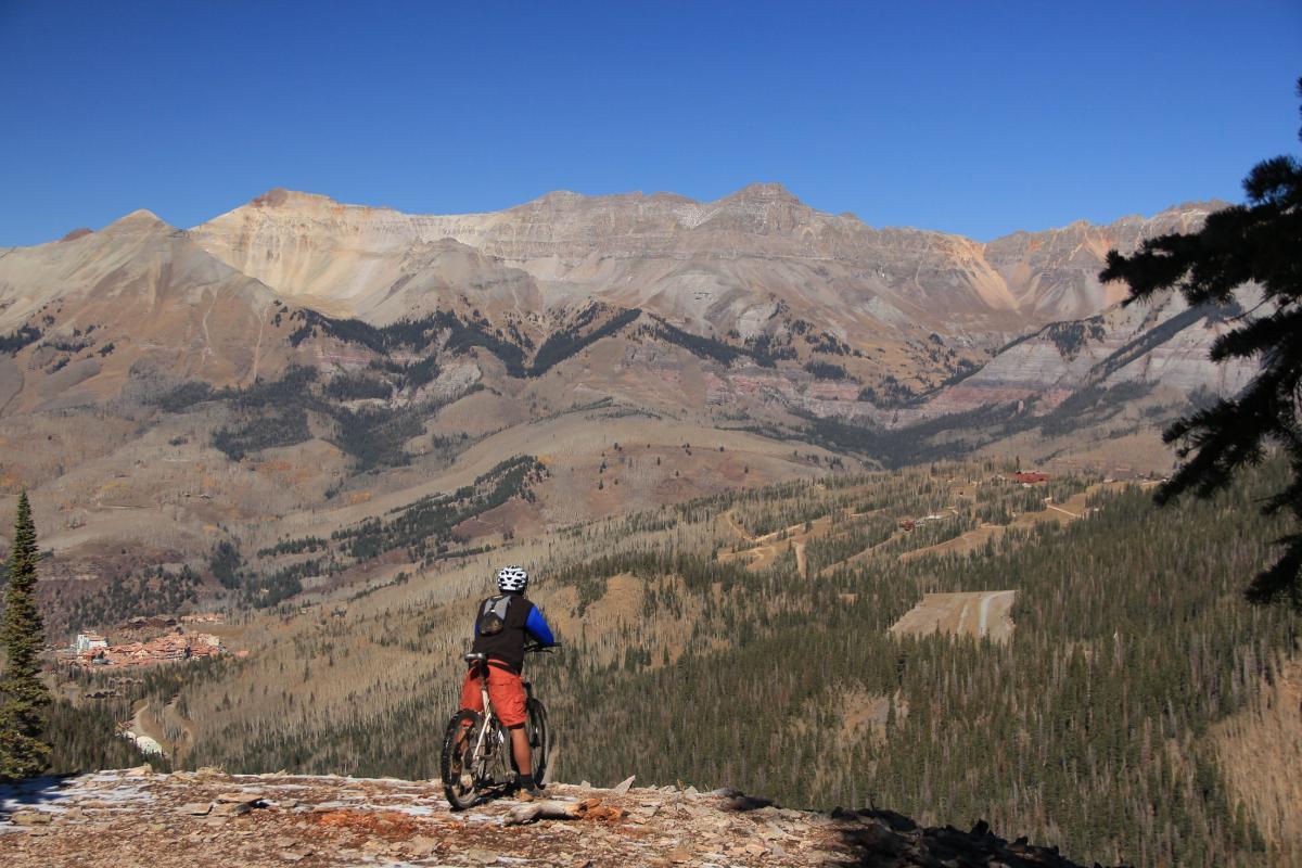 A mountain biker stands on a rocky outcrop overlooking a vast mountainous landscape, with rolling hills and dense pine forests below. The sky is clear and blue, and the sunlight highlights the rugged terrain of the mountains in the background. Prospect Trail mountain bike trail.