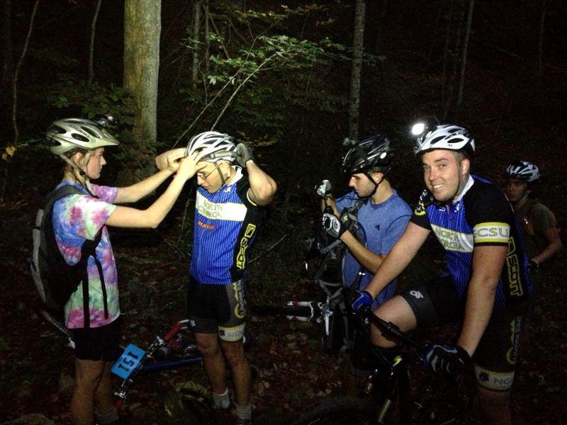 A group of mountain bikers adjusting their helmets in a wooded area at night. One person, wearing a colorful tie-dye shirt and a backpack, is helping another adjust their helmet while three other bikers are nearby, smiling and preparing their gear. The scene is illuminated by headlamp light, creating a lively atmosphere amidst the dark trees. Bull / Jake Mountain mountain bike trail.
