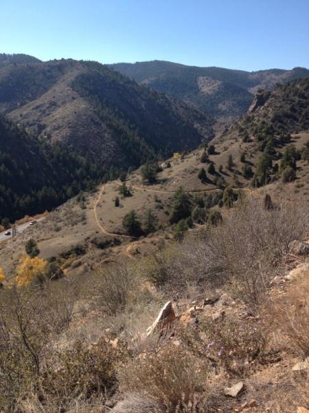 A scenic view of mountains and valleys under a clear blue sky, featuring rolling hills, sparse vegetation, and a dirt trail winding through the landscape. The foreground shows rocky terrain and scattered shrubs, while the background includes taller mountains, creating a picturesque natural environment. Centennial Cone Park mountain bike trail.