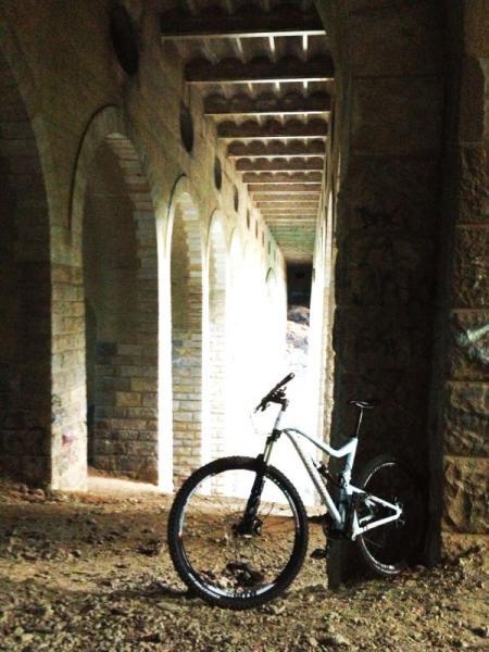 A mountain bike leaned against a stone pillar in an archway, surrounded by a tunnel-like structure with multiple stone arches. The scene features dim lighting, with light visible at the far end of the corridor, creating a contrasting atmosphere of shadows and illumination. Wissahickon Valley Park mountain bike trail.