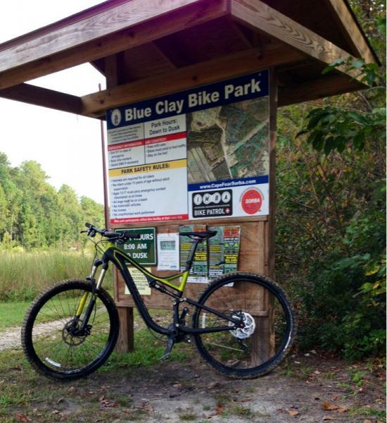 A mountain bike parked in front of a sign at the entrance of Blue Clay Bike Park, surrounded by greenery. The sign displays park hours, safety rules, and a map of the biking trails. Blue Clay Bike Park mountain bike trail.