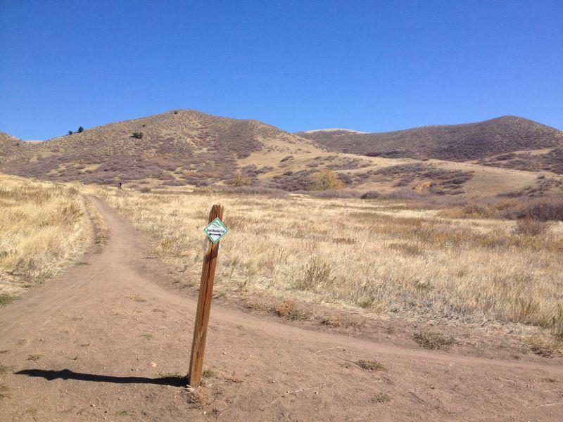 A dirt path forks in a field with sparse vegetation, leading towards rolling hills under a clear blue sky. A wooden signpost stands to the side of the path, indicating trail directions. Green Mountain mountain bike trail.