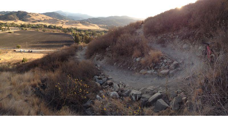 A scenic view of a winding dirt bike trail surrounded by dry, rocky terrain and sparse vegetation, with rolling hills and mountains in the background. The warm light of sunset casts a golden hue over the landscape. A bicycle is positioned on the right side of the image, partially obscured by the brush. Santa Teresa Park mountain bike trail.