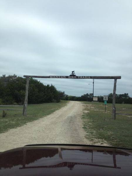 A dirt road leading into a mountain bike trail entrance, marked by a wooden archway with the text "MOUNTAIN BIKE TRAILS" at the top. The scene is set under a cloudy sky, with green foliage visible on either side of the path. Blora mountain bike trail.