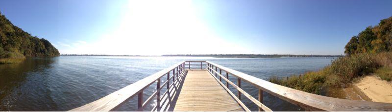 **Alt text:** A panoramic view of a wooden dock extending into a calm body of water, surrounded by lush greenery and shoreline, under a clear blue sky. Hartshorne Woods Park mountain bike trail.