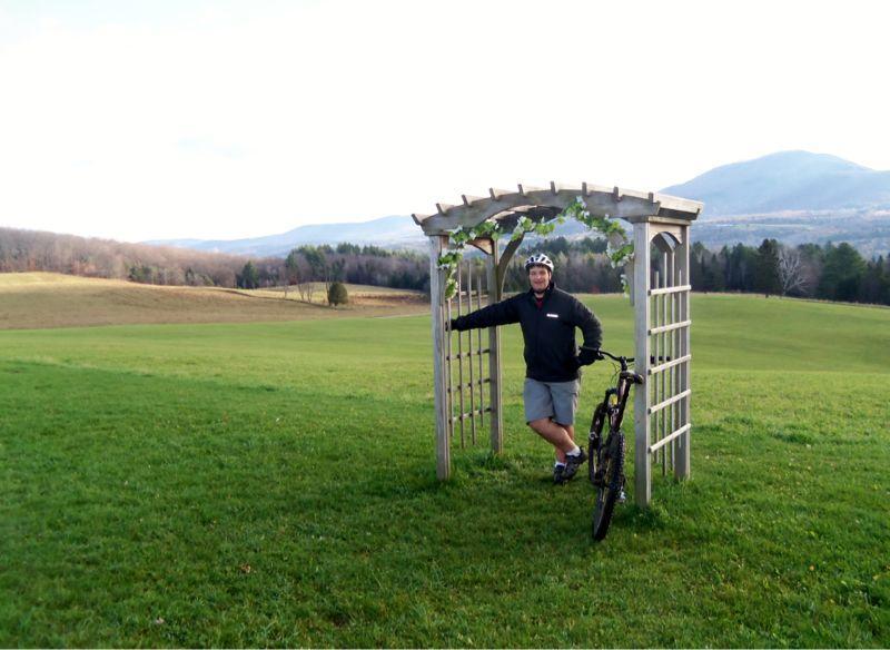 A man in a cycling outfit stands beside a mountain bike, leaning against a wooden arbor adorned with vines. He is positioned on a grassy field with rolling hills and mountains in the background under a partly cloudy sky. Kingdom Trails mountain bike trail.