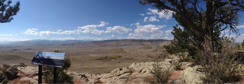 Panoramic view of a vast open landscape with rolling hills and a clear blue sky, featuring scattered clouds. In the foreground, there is a metal sign on a post, and the area is surrounded by rocky terrain and sparse vegetation, including pine trees. Rabbit Mountain Loop mountain bike trail.