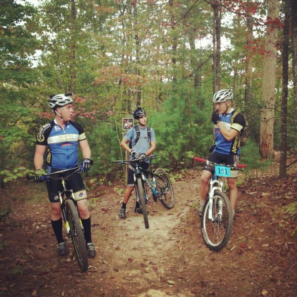 Three mountain bikers standing at a trail intersection in a wooded area. The first rider is wearing a blue and black jersey, black shorts, and has a serious expression. The second rider, dressed in a gray shirt and shorts, appears engaged in conversation. The third rider, also in a blue and black jersey and wearing bib number 151, is seen thinking as he holds the handlebars. Lush trees and greenery surround the path, indicating an autumn setting with hints of color in the leaves. Bull / Jake Mountain mountain bike trail.