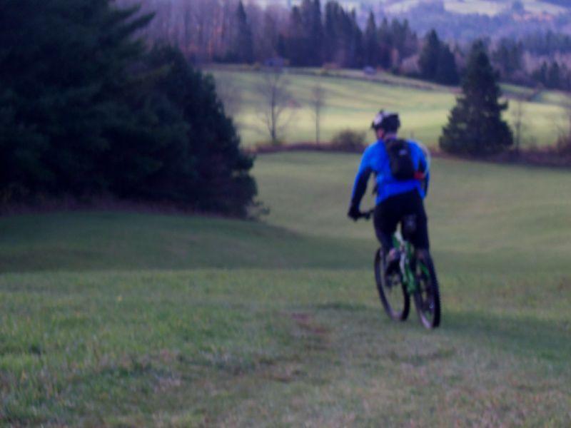 A person biking down a grassy hill, wearing a blue jacket and black pants, with a backpack. The background features a scenic landscape of trees and rolling hills. The image is slightly blurred, emphasizing movement. Kingdom Trails mountain bike trail.
