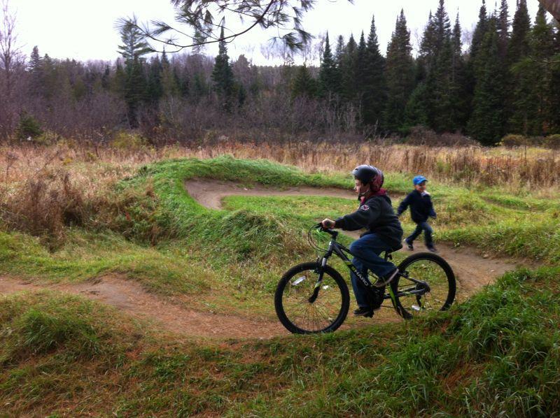 A child in a helmet rides a mountain bike on a dirt path surrounded by grassy mounds, while another child runs nearby in a forested area with tall trees and autumn foliage. Kingdom Trails mountain bike trail.