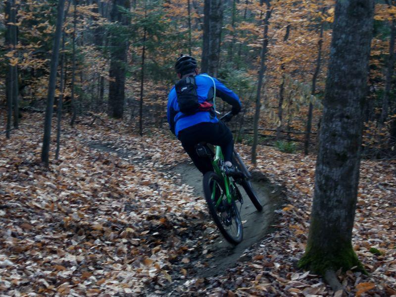 A mountain biker in a blue jacket rides along a winding, narrow dirt trail surrounded by autumn foliage, with fallen leaves covering the ground. Trees line the path, and the scene captures the essence of outdoor adventure in a forested setting. Kingdom Trails mountain bike trail.