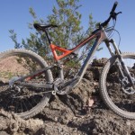 A mountain bike positioned on rocky terrain with sparse vegetation in the background, featuring a sleek frame with a combination of black and red colors. The bike's front wheel is turned slightly, showcasing its suspension and tire tread. The backdrop includes a clear blue sky and rugged landscape.
