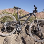 A mountain bike resting on rocky terrain, with rugged red hills and sparse vegetation in the background. The bike has a black frame and knobby tires, indicating it is designed for off-road use. The sky is clear and blue, suggesting a sunny day.