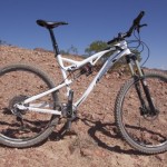A white mountain bike resting on rocky terrain under a clear blue sky, showcasing its rugged tires and suspension system. The backdrop features sparse vegetation typical of a desert environment.
