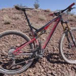A mountain bike resting on rocky terrain, showcasing a mix of black and red colors. The bike features thick tires designed for off-road trails, with visible components like the suspension forks and gears. Sparse vegetation is present in the background under a clear blue sky.