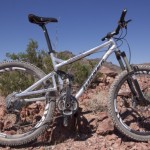 A silver mountain bike with large tires is leaning against rocky terrain, surrounded by desert vegetation. The backdrop features rugged mountains under a clear blue sky.