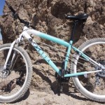 A mountain bike with a turquoise and white frame is leaning against a rocky outcrop in a desert landscape, under a clear blue sky. The bike features white tires and visible gears, showcasing its rugged design for outdoor adventures.