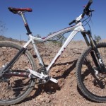 A mountain bike with a white and black frame, equipped with thick tires and a front suspension, parked on rocky terrain under a clear blue sky. Sparse vegetation is visible in the background.