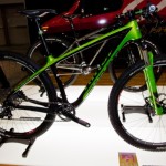 A modern mountain bike displayed on a stand, featuring a sleek black and neon green design. The bike has thin tires, dual suspension, and a minimalist frame, highlighting its lightweight construction. The background includes blurred figures and another bike, indicating a biking exhibition setting.
