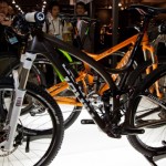 Two mountain bikes displayed in a showroom. The front bike is black with white accents, featuring a prominent front suspension fork. The second bike is orange with black and green highlights. In the background, a few people can be seen observing the bikes, adding to the lively atmosphere of the exhibition.