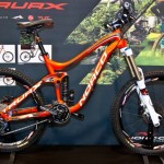 Image of a bright orange mountain bike displayed on a black stand. The bike has wide tires, a sleek frame, and features designed for off-road performance. It is set against a backdrop showcasing outdoor trails and mountain biking visuals.