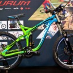 A brightly colored mountain bike with a green and blue frame on display at an event, featuring wide tires and a prominent suspension system. Behind it, there is a promotional backdrop with images and logos related to biking.