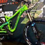 A vibrant green mountain bike displayed at an exhibition, featuring a prominent front suspension fork and specialized tires designed for rugged terrain. The background includes promotional graphics related to biking, showcasing scenic trails and the bike's brand logo.