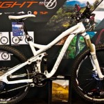 A mountain bike featuring a sleek, white frame with prominent suspension and wide tires, displayed against a promotional backdrop showcasing outdoor landscapes and cycling activities.