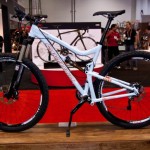 Mountain bike displayed at a cycling expo, featuring a lightweight white frame and prominent suspension system. The bike is positioned on a wooden stand with a red carpet background, surrounded by various exhibitors and attendees.