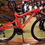 A vibrant red mountain bike displayed on a wooden stand, showcasing its features and design at a trade show. In the background, attendees are engaged with their phones and other bikes.