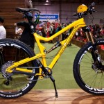 A bright yellow mountain bike displayed on a wooden platform, featuring high-performance components and large tires with white lettering. In the background, several people can be seen, along with various biking gear and accessories in a trade show setting.
