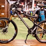 A high-performance mountain bike displayed on a wooden stand at an event, featuring a sleek black frame, high-quality suspension forks, and distinctive wheels with branding. The background shows blurred figures and other bikes, indicating a busy show environment.