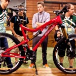 A vibrant red mountain bike featuring high-performance components is displayed prominently on a wooden stand. Three individuals, engaged in conversation, examine the bike. The background consists of a rustic wooden wall, creating an inviting atmosphere for a cycling event. The scene captures the excitement of showcasing new biking technology.