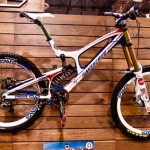 A high-performance mountain bike displayed on a wooden wall. The bike features a sleek design with a white frame accented by blue and red graphics. It has prominent front suspension forks, a multi-gear drivetrain, and large, rugged tires suitable for off-road terrain.