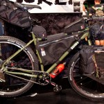 A green mountain bike equipped with several black storage bags for bikepacking, showcasing a rugged design. The bike is displayed on a black surface with blurred backgrounds of a trade show or event, emphasizing its adventure-ready features.