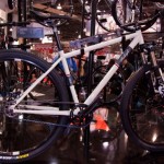 A white mountain bike displayed at an indoor exhibition, showcasing its sleek frame, thick tires, and front suspension. The bike is positioned on a glossy black surface, with other bicycles and display elements in the background.
