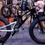 A black and white fat bike with oversized tires displayed at a cycling exhibition, with attendees in the background examining other bikes. The bike features a modern design and is equipped with front suspension.