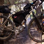 A mountain bike displayed in a booth at a trade show, featuring a lightweight aluminum frame and various accessories, including a frame bag and a seat bag, set against a backdrop of trees and a dirt path. The bike has knobby tires and appears to be designed for off-road riding, with dirt visible on the bike's components indicating previous use.