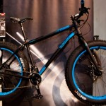 A black and blue mountain bike is displayed against a dark backdrop, showcasing its large, rugged tires and sleek frame design. The bike features bold blue accents on the wheels and frame, highlighting its sporty aesthetic.
