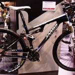 Alt text: A black and white mountain bike with a sleek design, featuring a prominent "Jamis" logo. The bike is displayed on a stand at a trade show. In the background, blurred figures can be seen, indicating a busy indoor event.