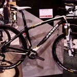 A black and white mountain bike displayed on a stand, showcasing its features including a lightweight frame, front suspension fork, and sleek design. A price tag is visible, indicating a discount. The background hints at an exhibition setting with other bikes and attendees.